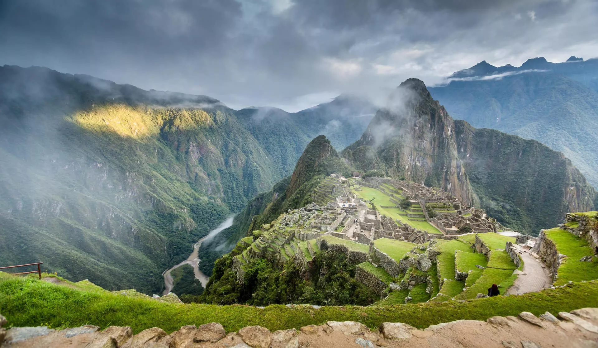 Vista panorámica de las ruinas de piedra de Machu Picchu y terrazas verdes bajo un espectacular cielo nublado con la luz del sol atravesando la niebla.