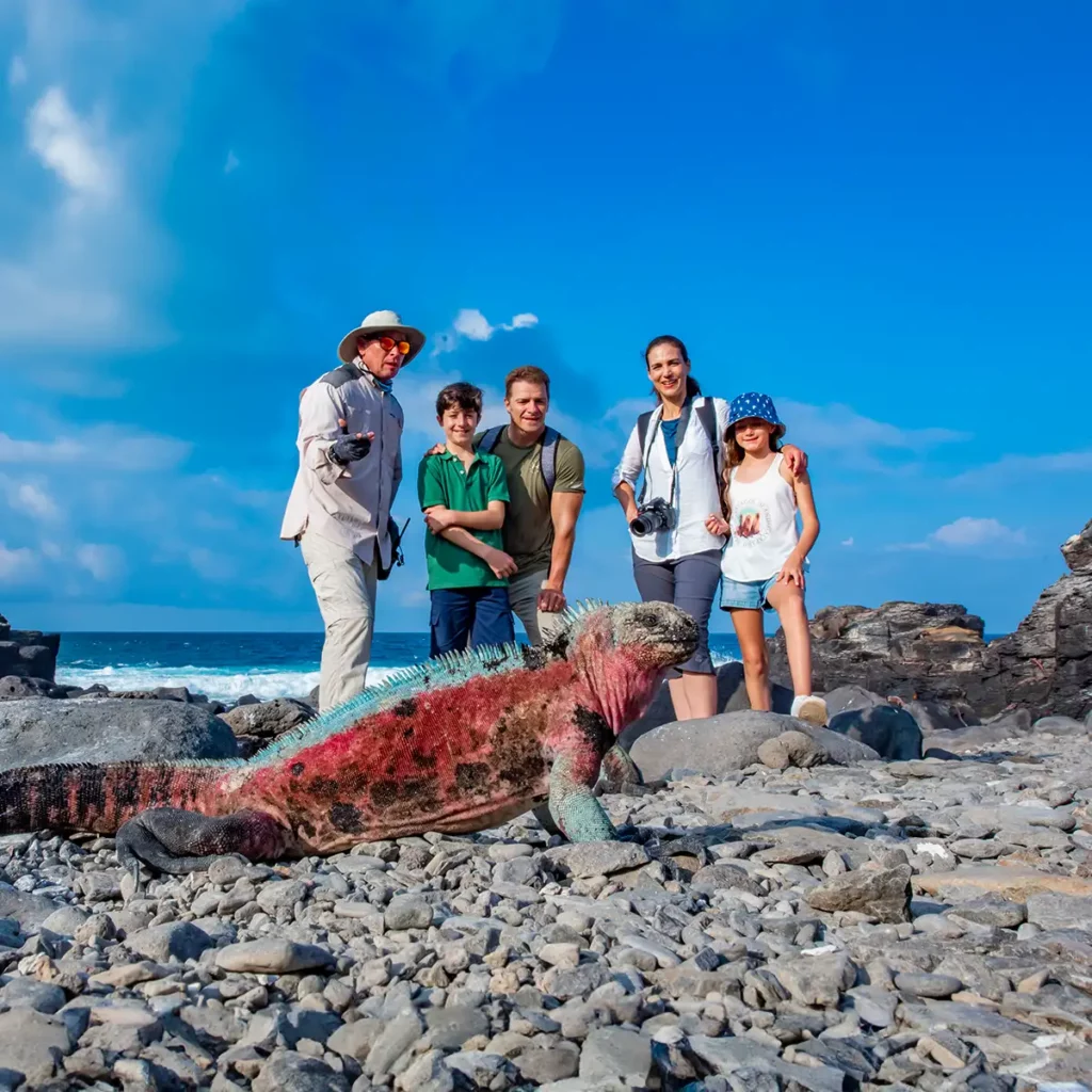 Familia en una costa rocosa de Galápagos observando una iguana marina con el océano al fondo.
