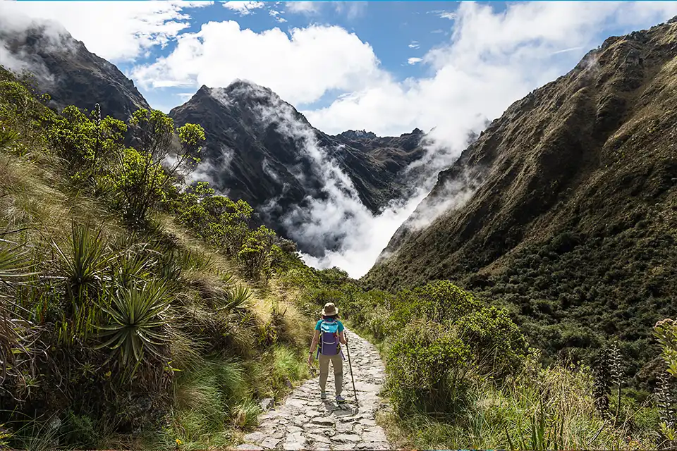 Una excursionista con una mochila y un bastón de trekking recorre un sendero de piedra durante la mejor época para recorrer el Camino Inca, rodeada de brumosas montañas andinas y exuberante vegetación.