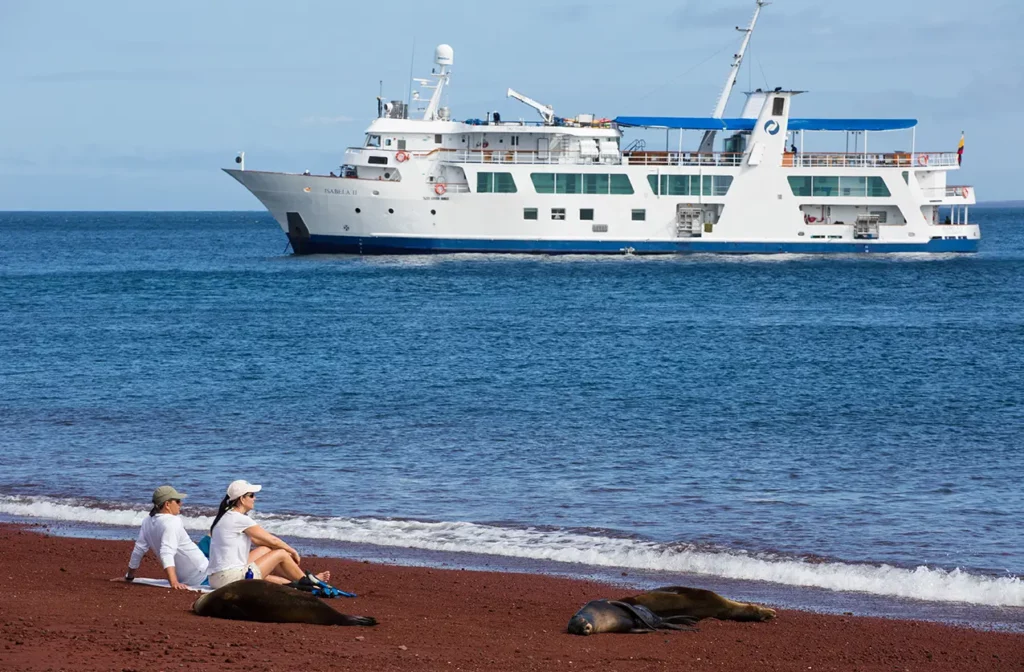 Two guests relaxing on red sand beach beside sea lions with Yacht Isabela II anchored offshore Galapagos